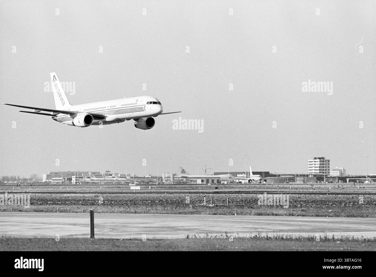 New Boeing 757 from Transavia Schiphol, Whizgle News, Dutch Desk, The Netherlands, 1950 - 2000 on 22-02-1993. The image shows these topics. In the scene, a commercial aircraft is captured in mid-flight, approaching its landing. The plane is predominantly white with subtle markings, featuring a streamlined shape characteristic of modern airliners. Its wings are outstretched, and the landing gear is partially deployed, indicating a descent. The backdrop consists of an expansive airport, with multiple terminal buildings visible in the distance. The architecture is functional and modern, character Stock Photo