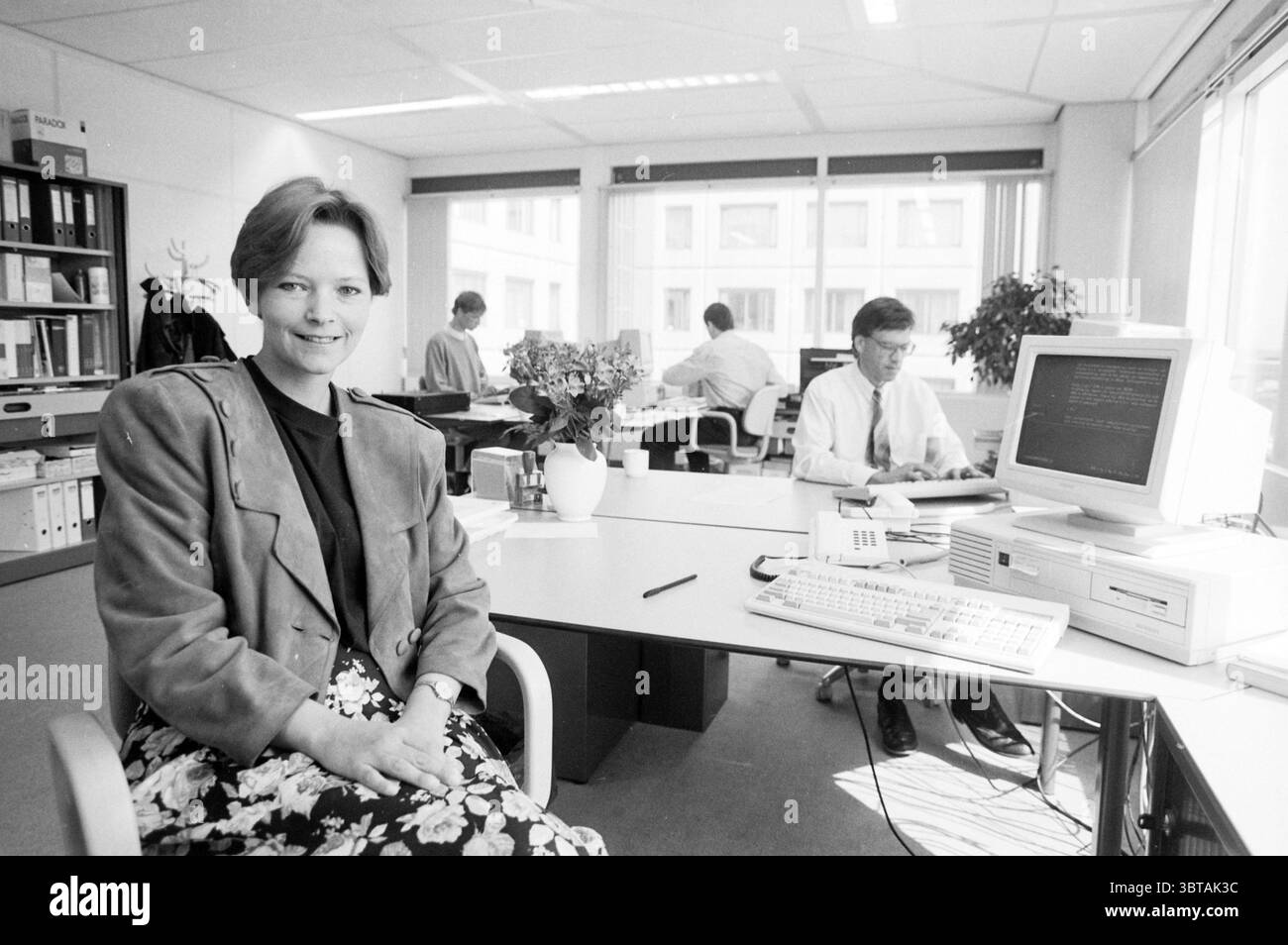 Mrs. Trel MKA H'dorp Hoofddorp The Netherlands, Whizgle News, Dutch Desk, The Netherlands, 1950 - 2000 on 18-04-1991. The image includes these topics. The scene is set in a modern office environment that conveys a sense of professionalism and focus. A woman sits confidently at a desk, her expression engaging and friendly. She is dressed in a stylish blazer and a floral skirt, which add a touch of personality to her appearance. The color palette features soft, muted tones that enhance the calm atmosphere of the space. In the background, a few colleagues are occupied with their work. One individ Stock Photo