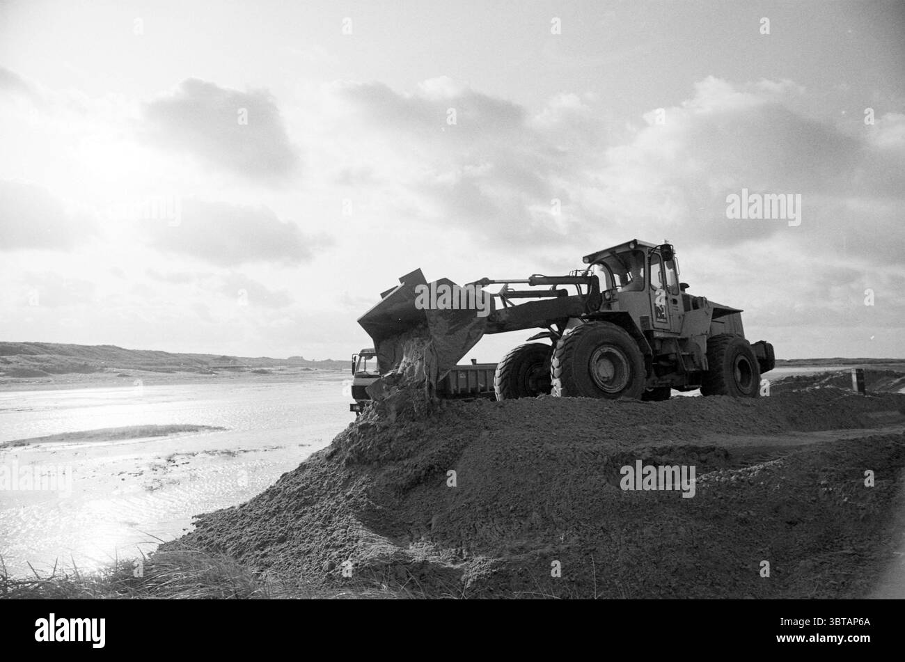 First beach team activities, Whizgle News, Dutch Desk, The Netherlands, 1950 - 2000 on 17-11-1992. These topics are shown in the image. The scene captures a working environment characterized by a monochromatic palette, where various shades of gray dominate. A large earth-moving vehicle, possibly a front loader, is positioned prominently in the foreground. Its mechanical arm is raised high, poised mid-action as it scoops up a mound of soil, sending a cascade of dirt tumbling down its side. The ground in the area appears uneven and composed of freshly disturbed earth, indicating recent excavatio Stock Photo