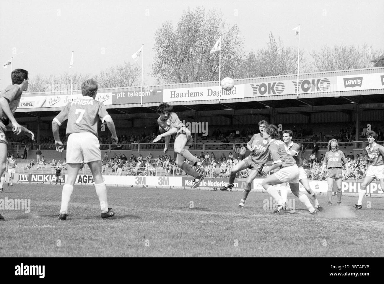 Haarlem - AZ, Whizgle News, Dutch Desk, The Netherlands, 1950 - 2000 on 13-04-1991. These topics appear in the image. The scene depicts an intense soccer match unfolding on a grass field under bright daylight. A group of players in vibrant jerseys, predominantly orange, are engaged in dynamic motion; one player is airborne, leaping with determination to head the ball. The expressions on the players’ faces convey focus and competitiveness. The other athletes are poised, some with arms outstretched and feet planted, while others are in motion, ready to react. The setting features a stadium fille Stock Photo