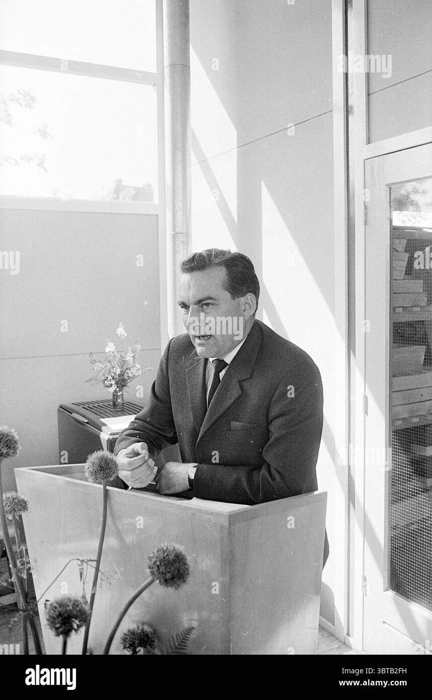 Opening Krelageschool Opening openings commissioning, Whizgle News, Dutch Desk, The Netherlands, 1950 - 2000 on 20-05-1966. These are the topics in the image. The scene features a man standing at a lectern made of light-colored wood, positioned in a modern interior space with large windows allowing natural light to spill in. The man is dressed in a dark suit, which contrasts sharply with the lighter tones of his surroundings. His expression is serious and animated, suggesting he is engaged in a passionate discussion or presentation. The background is minimalistic, highlighting the clean lines Stock Photo