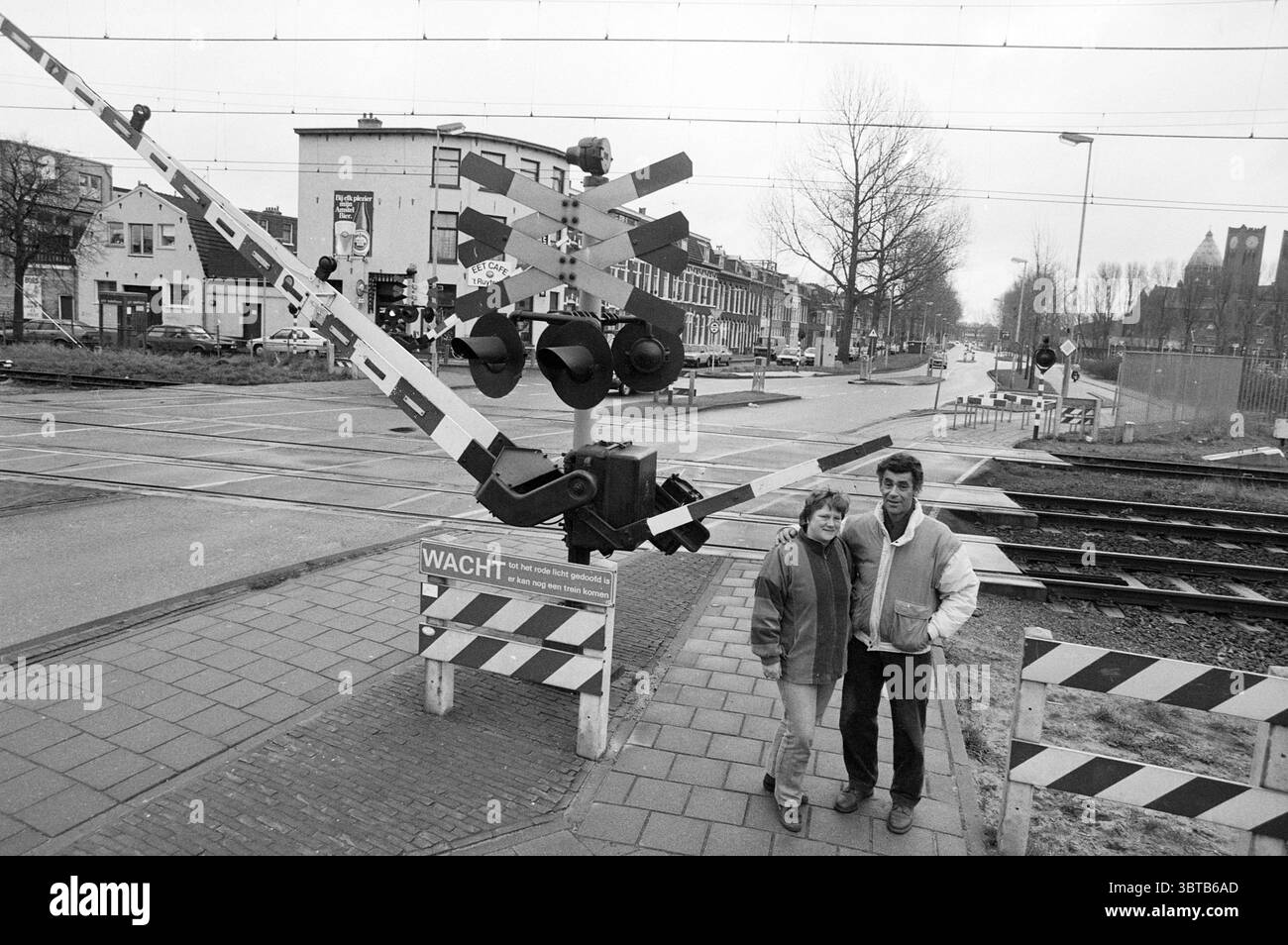 Two people at railway crossing Haarlem Westergracht Nederland, Whizgle News, Dutch Desk, The Netherlands, 1950 - 2000 in 1988. These topics appear in the image. The scene depicts two individuals standing in front of a level crossing, characterized by a stark, monochromatic palette. The foreground features a large, mechanical gate, angled downwards, adorned with bold striped patterns in black and white. This barrier creates a sense of caution, while the dim light of the day adds an overcast atmosphere. The two figures stand closely together, conveying a friendly yet contemplative mood. One pers Stock Photo