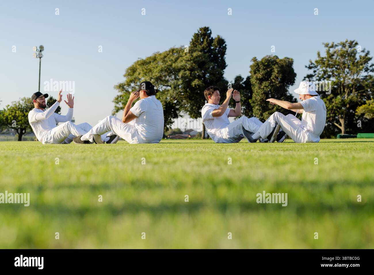 Diverse male teammates doing sit-ups and high-fiving on grass in cricket uniforms under floodlight Stock Photo