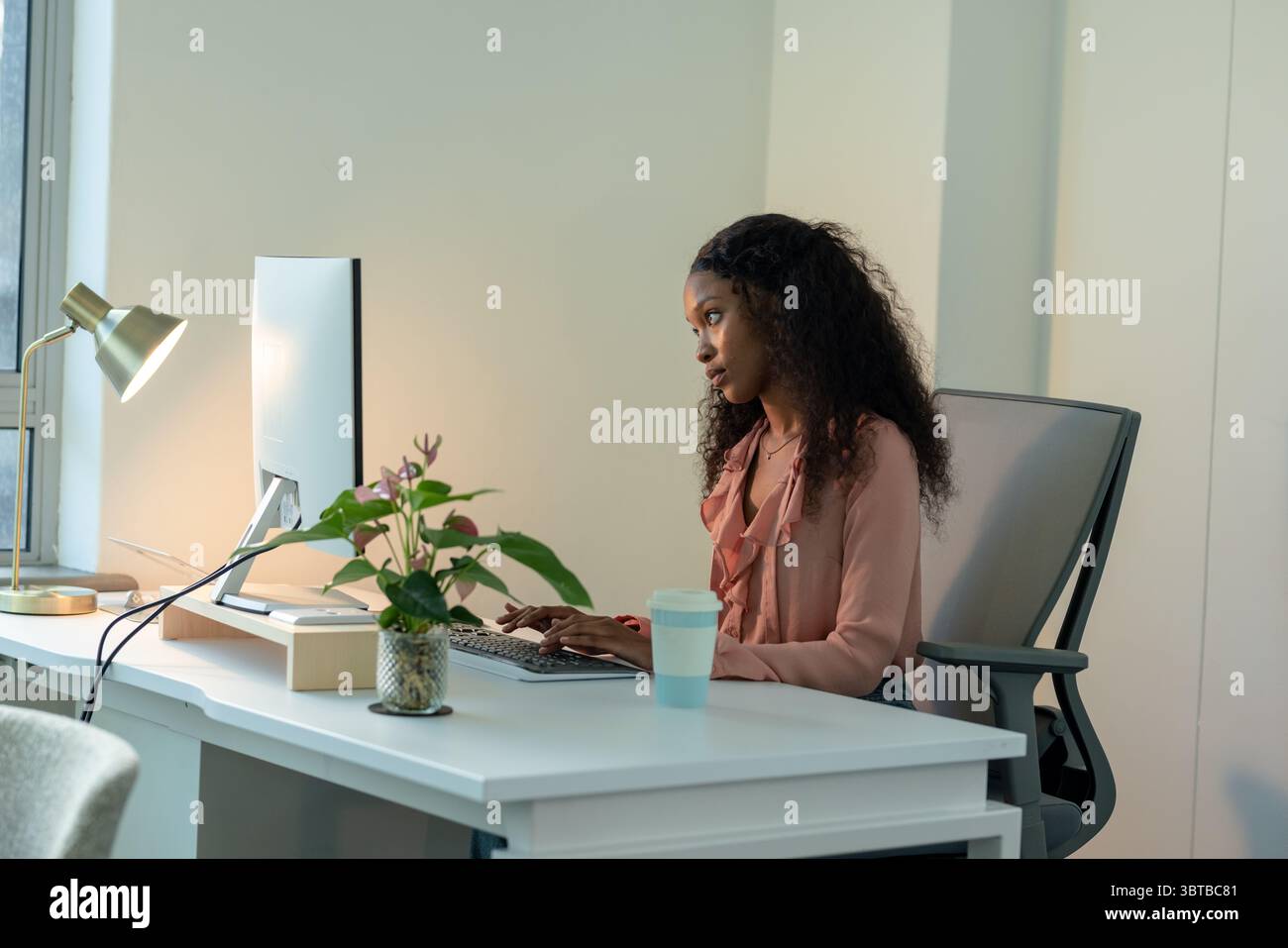 African American woman typing on keyboard at office desk with monitor and gold lamp, copy space Stock Photo