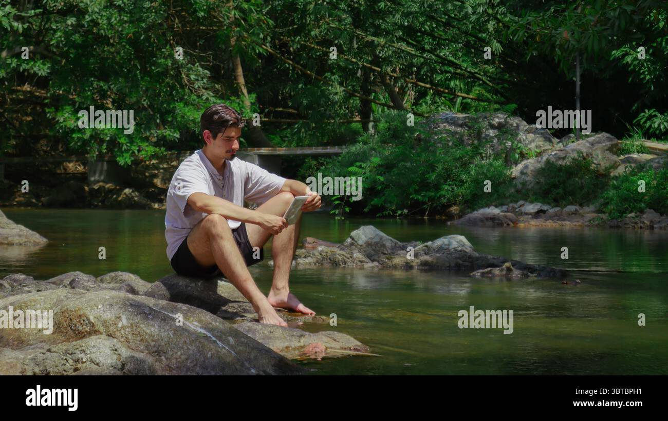 Young man sitting barefoot on rocks by a river in the jungle, reading a tablet, tropical forest scenery Stock Photo