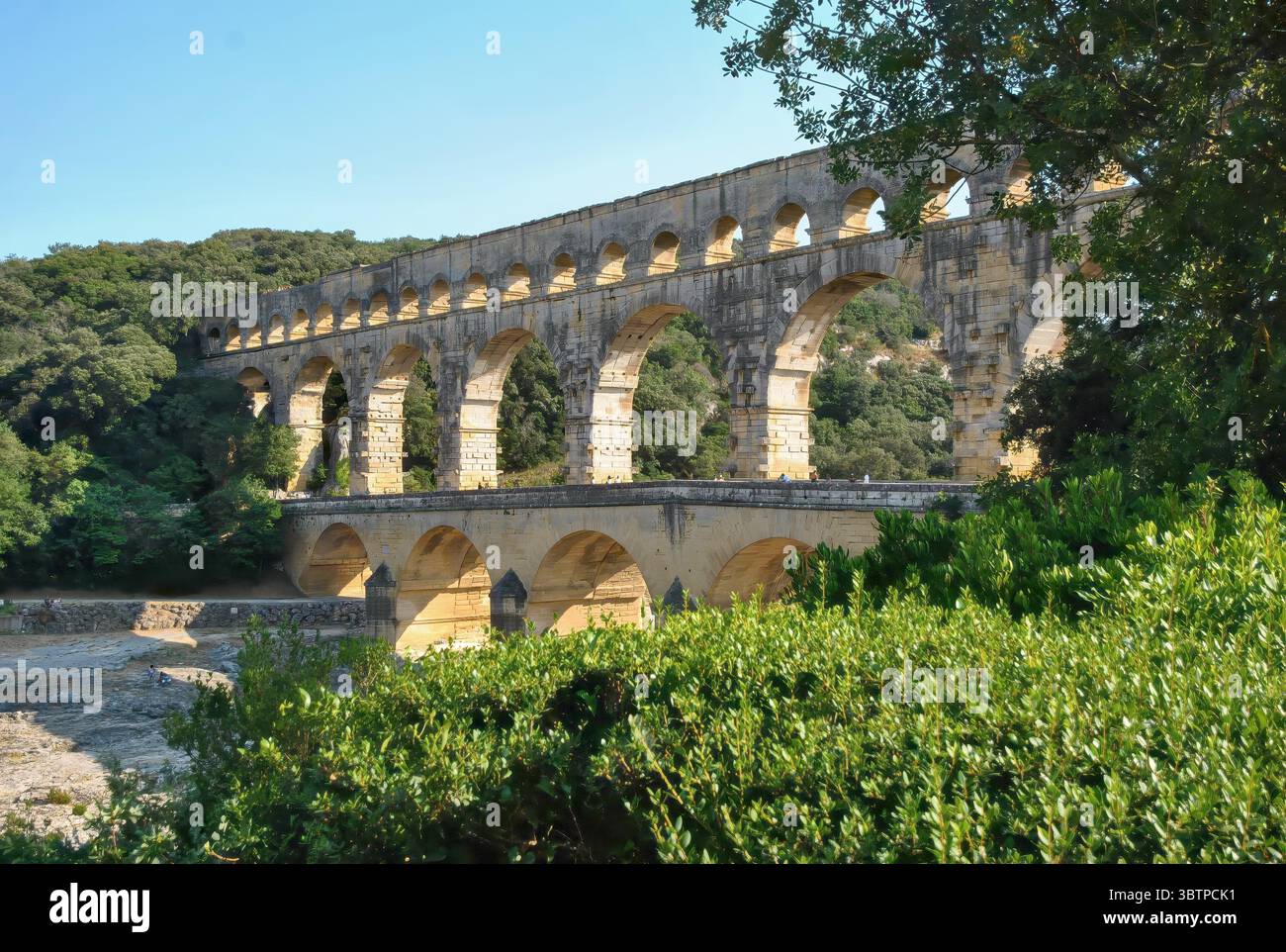 Ancient Roman aqueduct bridge crossing a river, surrounded by green forest under a clear blue sky. Stock Photo