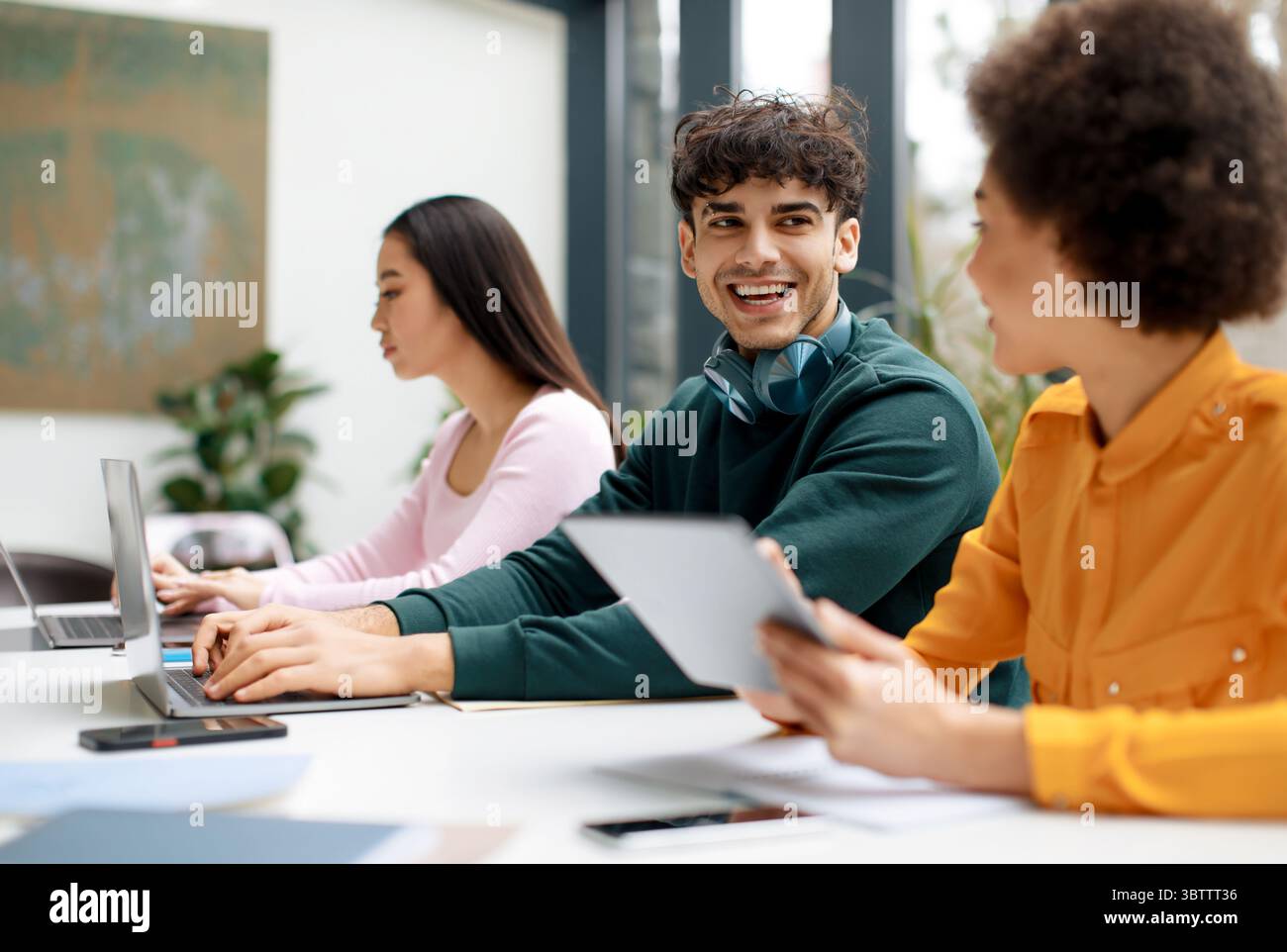 Happy diverse students sitting in classroom and communicating, using gadgets while being absorbed in studying Stock Photo
