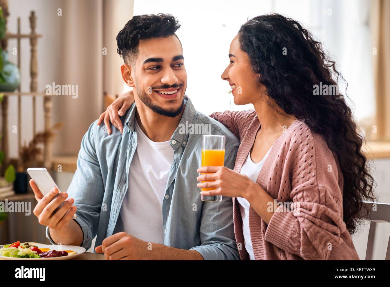Happy Young Arab Couple Using Smartphone During Breakfast In Kitchen Stock Photo