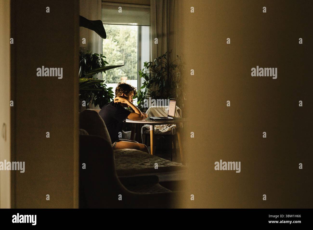 Teen sitting at table with laptop, studying or working at home, seen through doorway indoors Stock Photo