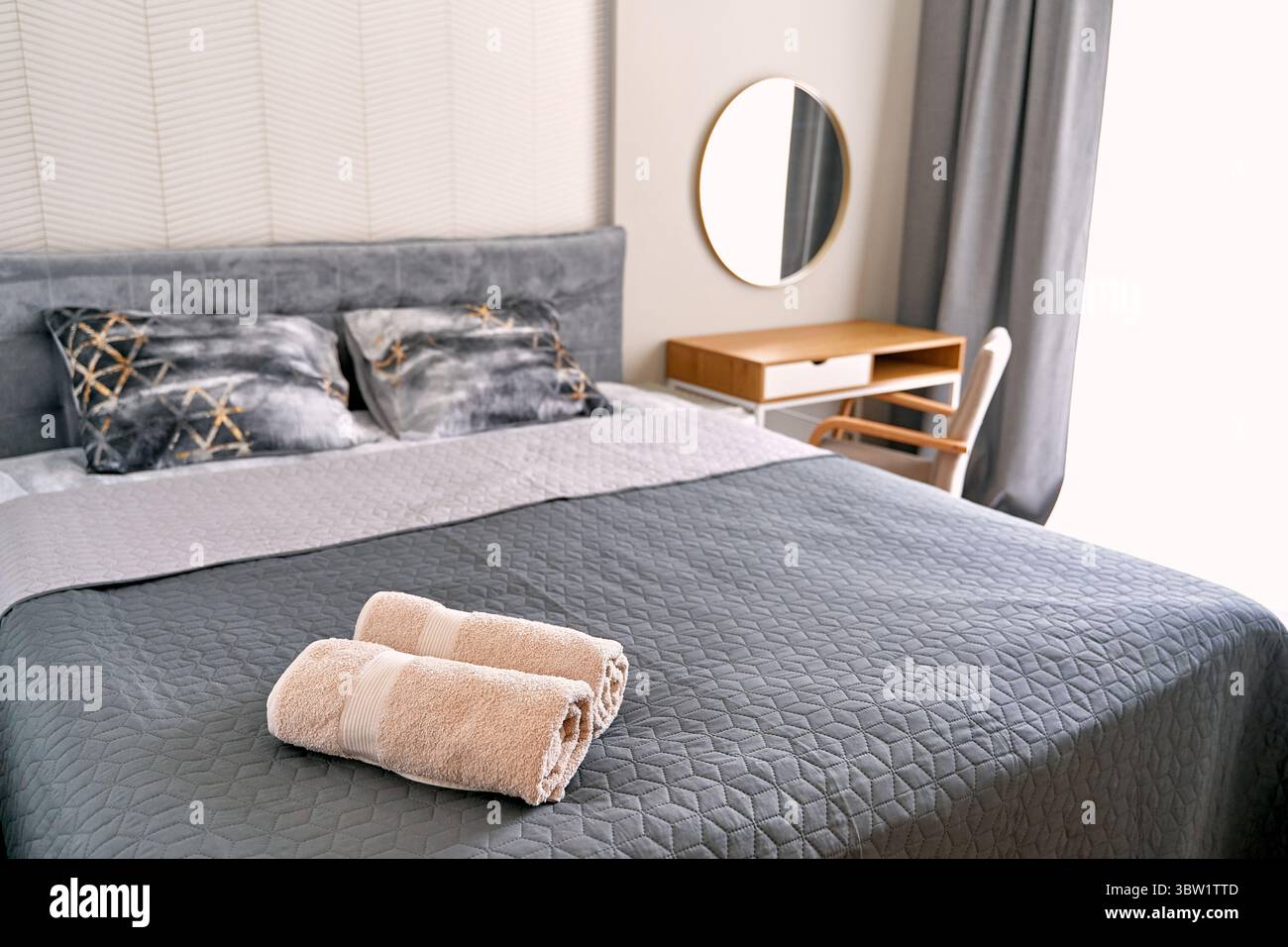 An elegant modern bedroom featuring gray bedding and decorative pillows on a comfortable bed, enhanced by a wooden desk, chair, and a stylish round mi Stock Photo