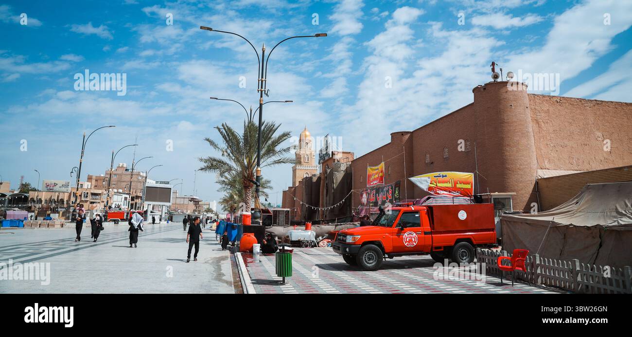 Bustling streets of Kufa showcase local shops, vehicles, and pedestrians enjoying a sunny day in Iraqs rich cultural backdrop. Stock Photo