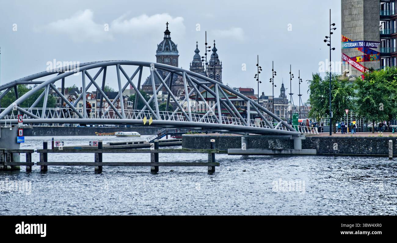 Modern arch bridge for bicycles crossing the amstel river, with the monumental saint nicholas basilica and typical dutch houses in the background, in Stock Photo