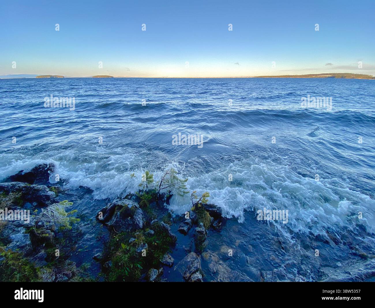 The ocean waves roll in, meeting a rugged, rocky shoreline under a blue sky. - Smartphone Captured Stock Image