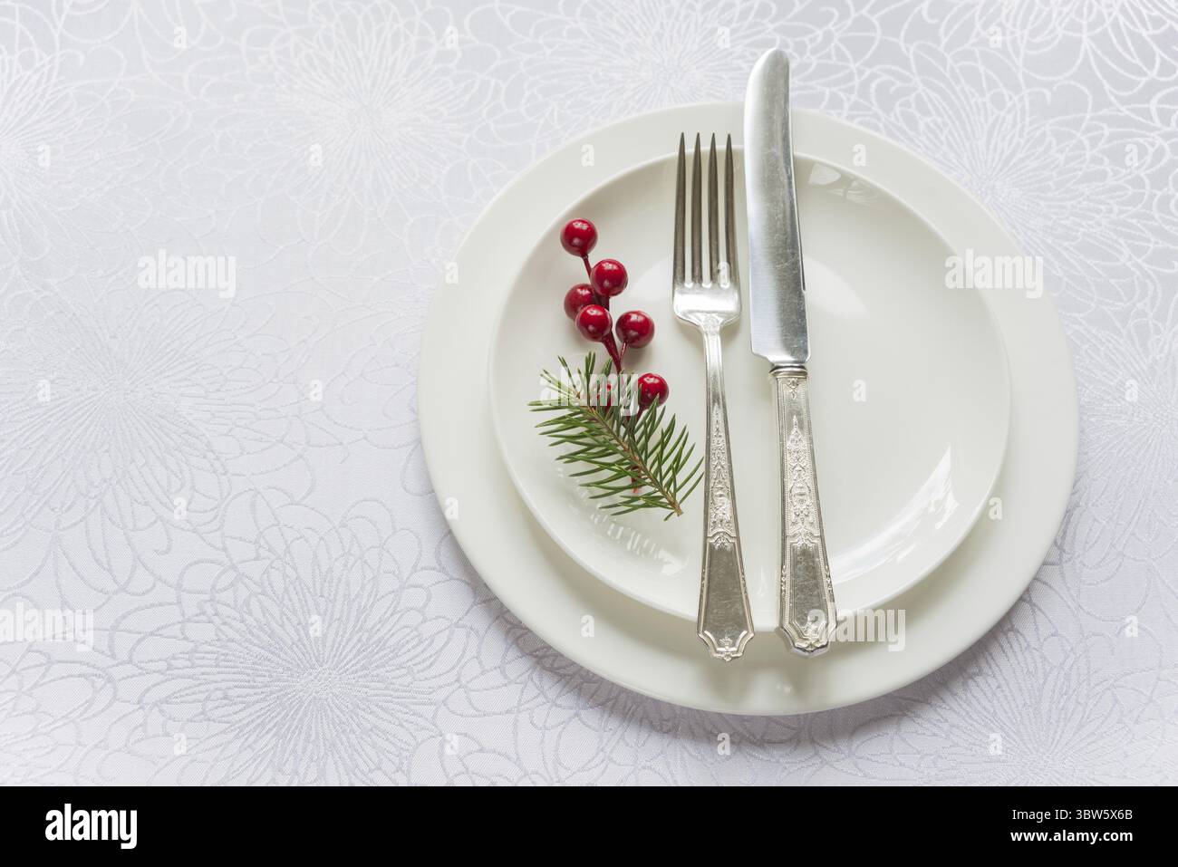 Christmas table in tones of white, beautifully served luxurious tableware: porcelain plates and silverware, decorated by fir sprig and holly berries, Stock Photo