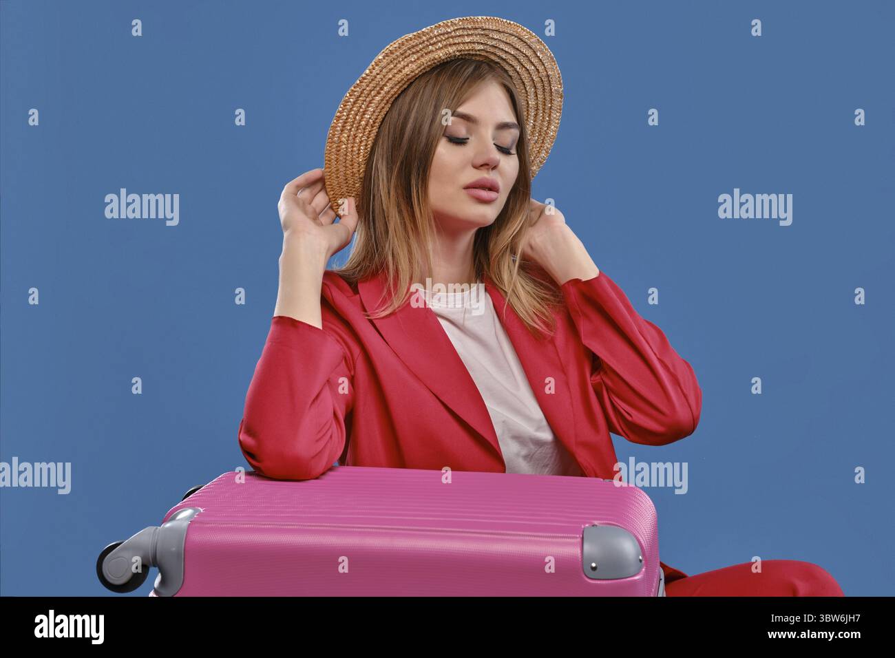 Charming blonde model in straw hat, white blouse, red pantsuit. She closed her eyes, touching headdress, sitting leaning on pink suitcase, posing on b Stock Photo