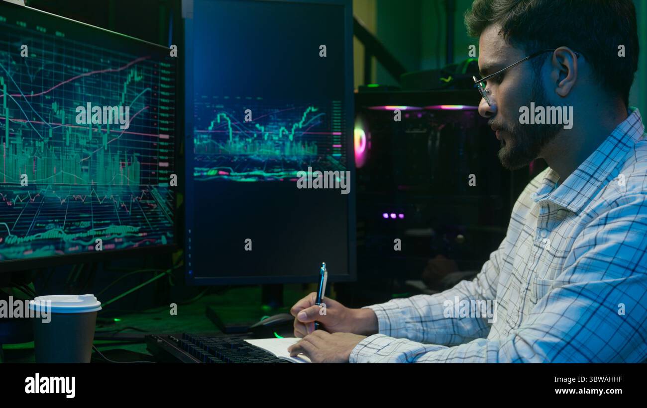 Indian Arabian man writing notes notebook working with pc computer trader programmer coder hacker male complete finish work online businessman night Stock Photo
