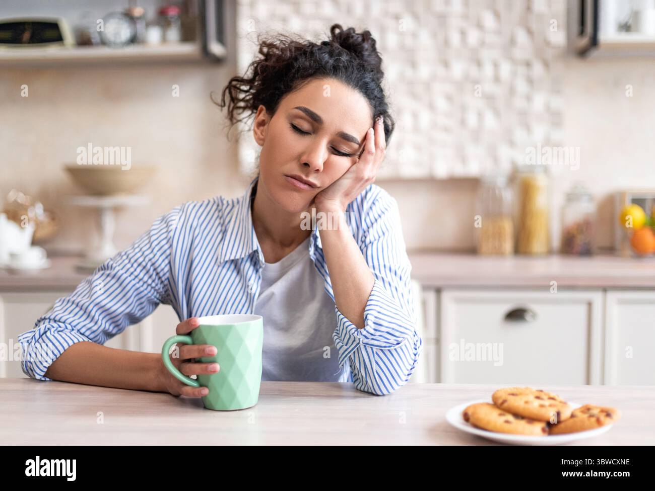 Sleepy young woman sitting at dining table in kitchen with closed eyes, holding cup while having breakfast Stock Photo