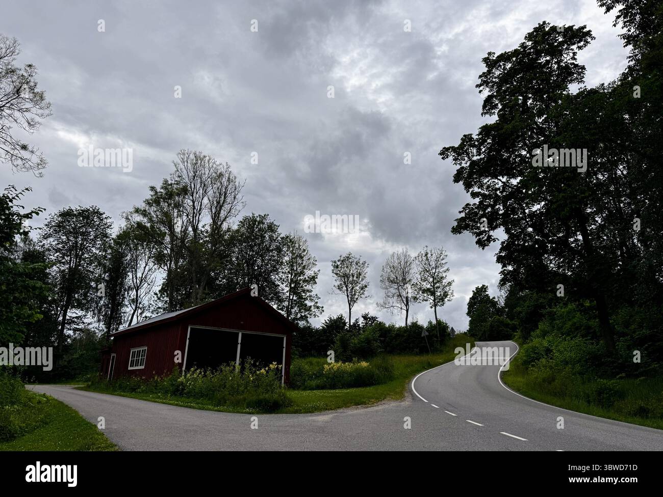 Curvy Country Road Under Moody Sky: Winding road and red barn in Fagervik, Inkoo, Uusimaa, Finland. A peaceful scene with strong historical atmosphere - Smartphone Captured Stock Image