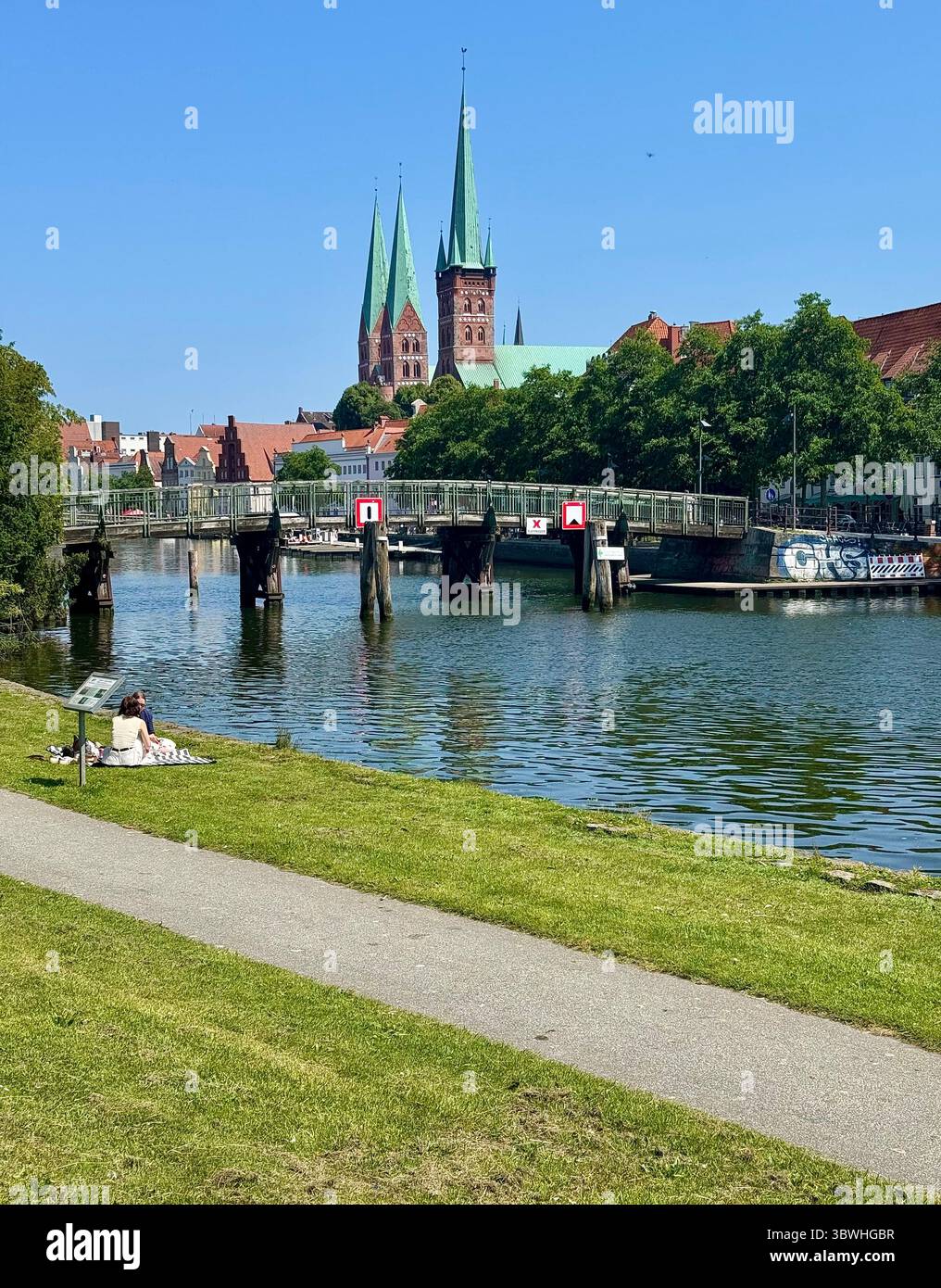 View of Lübeck’s old town with St. Mary's Church spires in the background, a riverside park in the foreground, and boats moored along the canal on a s - Smartphone Captured Stock Image