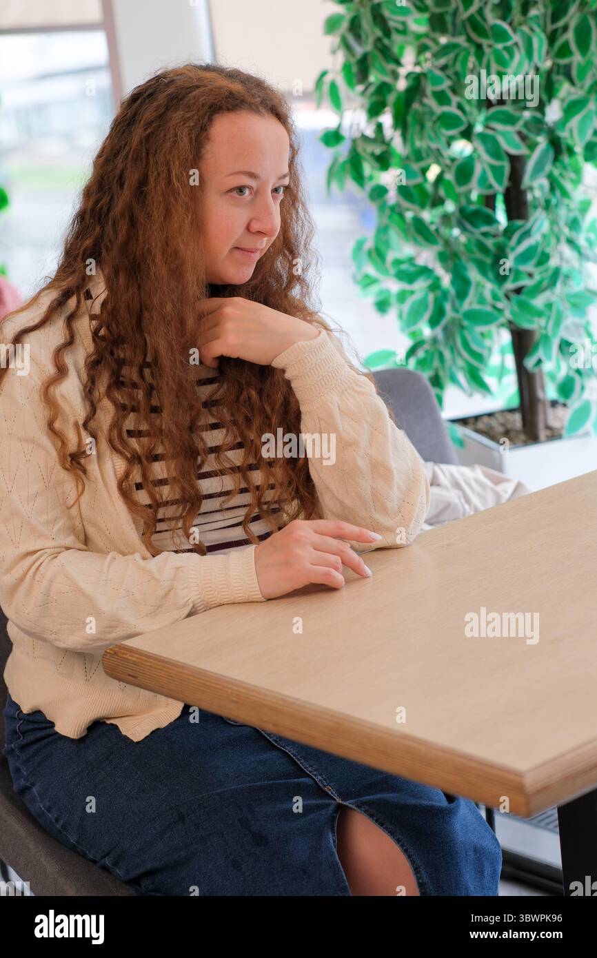 A young woman in a cozy sweater sits at a cafe table, deep in thought. Stock Photo