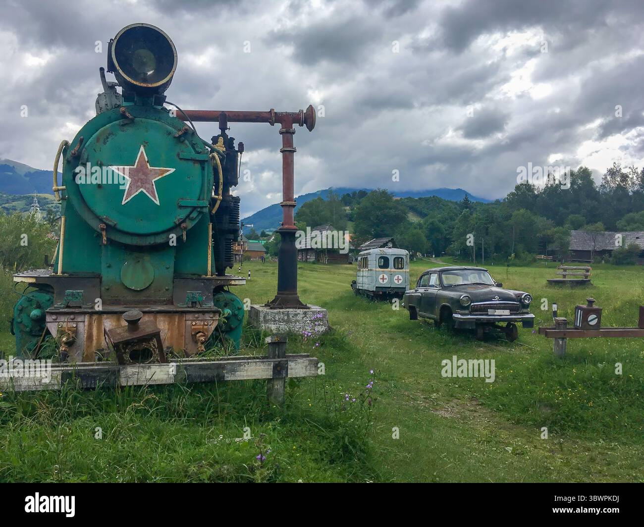 Old steam locomotive, vintage ambulance train and Soviet-era car on display in a rural open-air museum in Zakarpattia Oblast, Ukraine, under a cloudy Stock Photo