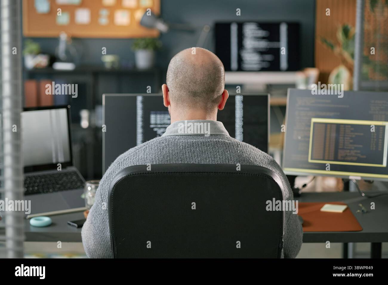 Middle aged Caucasian man sitting at desk working on multiple computer monitors with programming code visible on screens in modern office environment, back view Stock Photo