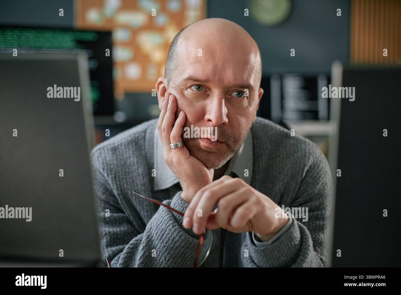 Portrait of middle aged Caucasian man resting chin on hand while sitting at desk, gazing intently at computer monitors, holding eyeglasses, appearing focused on work Stock Photo