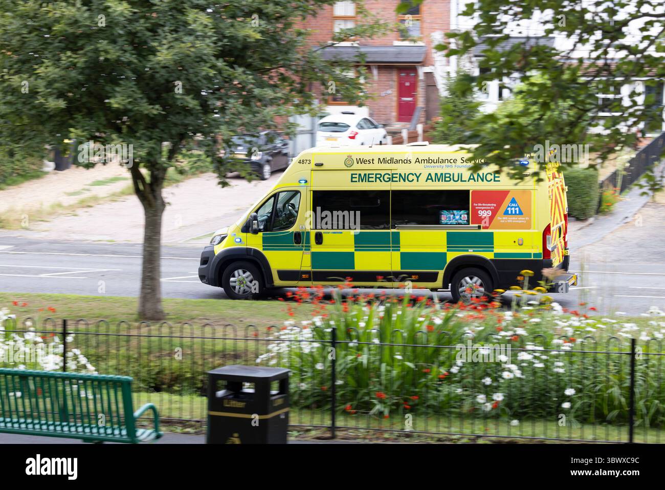 Side view of UK ambulance emergency vehicle travelling past a park with blue lights on heading to an emergency call. Stock Photo