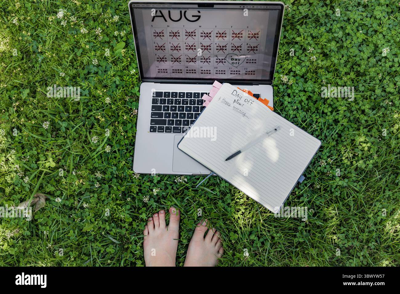 Feet in grass next to laptop and notebook with day off plan under natural light. Stock Photo