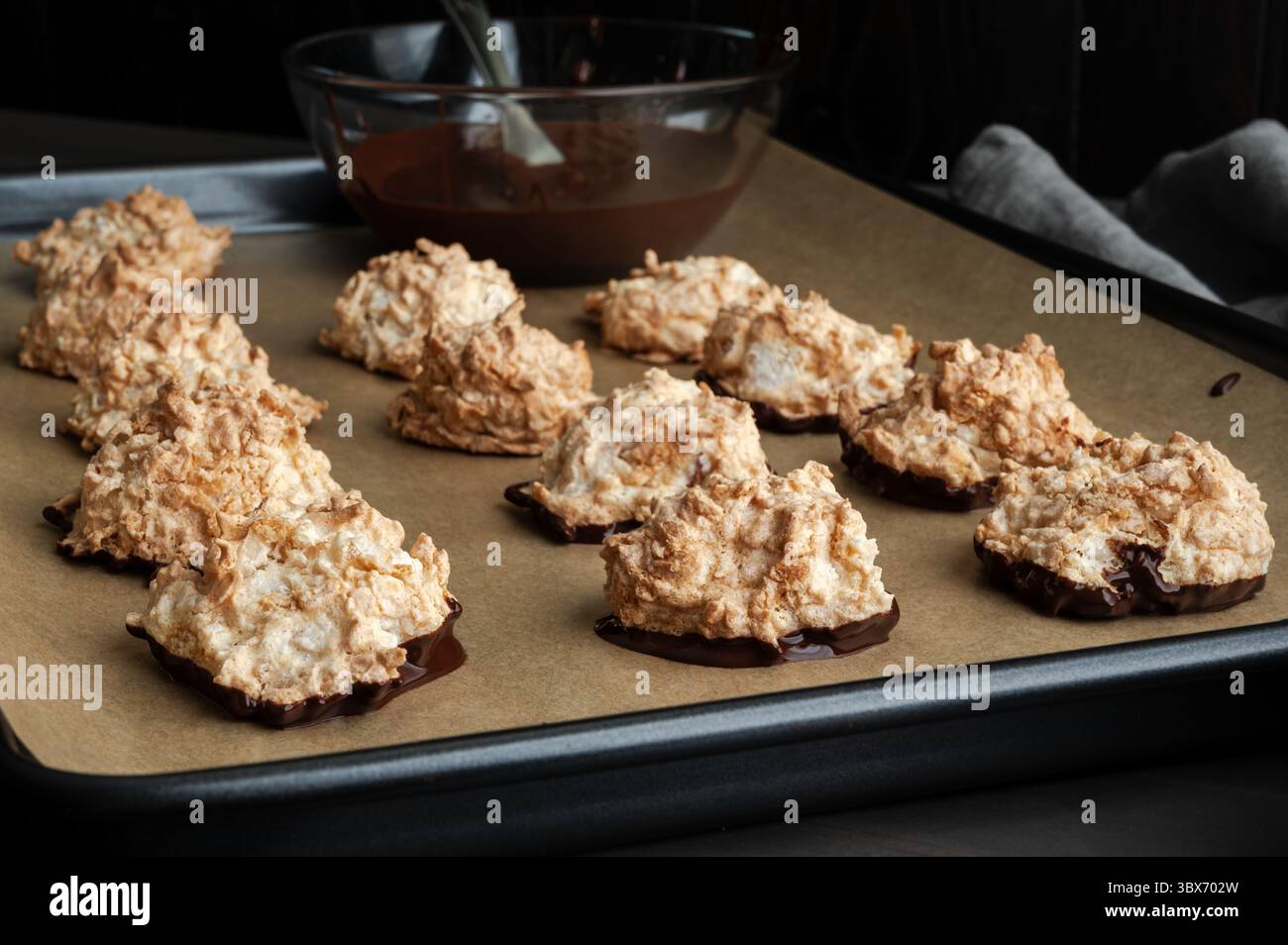 Macaroons Dipped in Dark Chocolate on a Sheet Pan: Coconut meringue drop cookies with a bowl of melted bittersweet chocolate on parchment paper Stock Photo