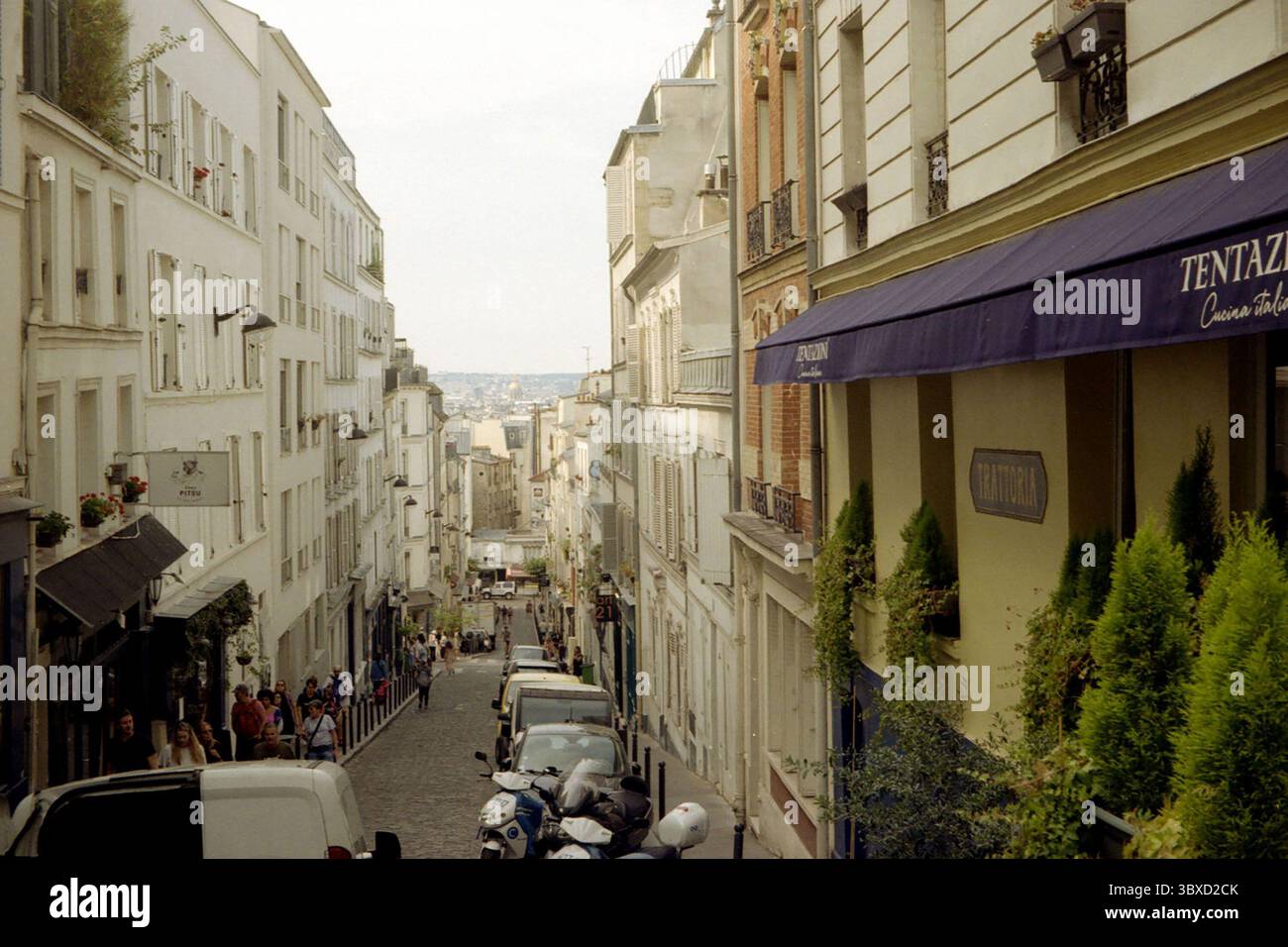 Candid summer street view on Rue Lepic in Montmartre, Paris, with pedestrians, parked cars, and restaurant awning, shot on 35mm film. Stock Photo