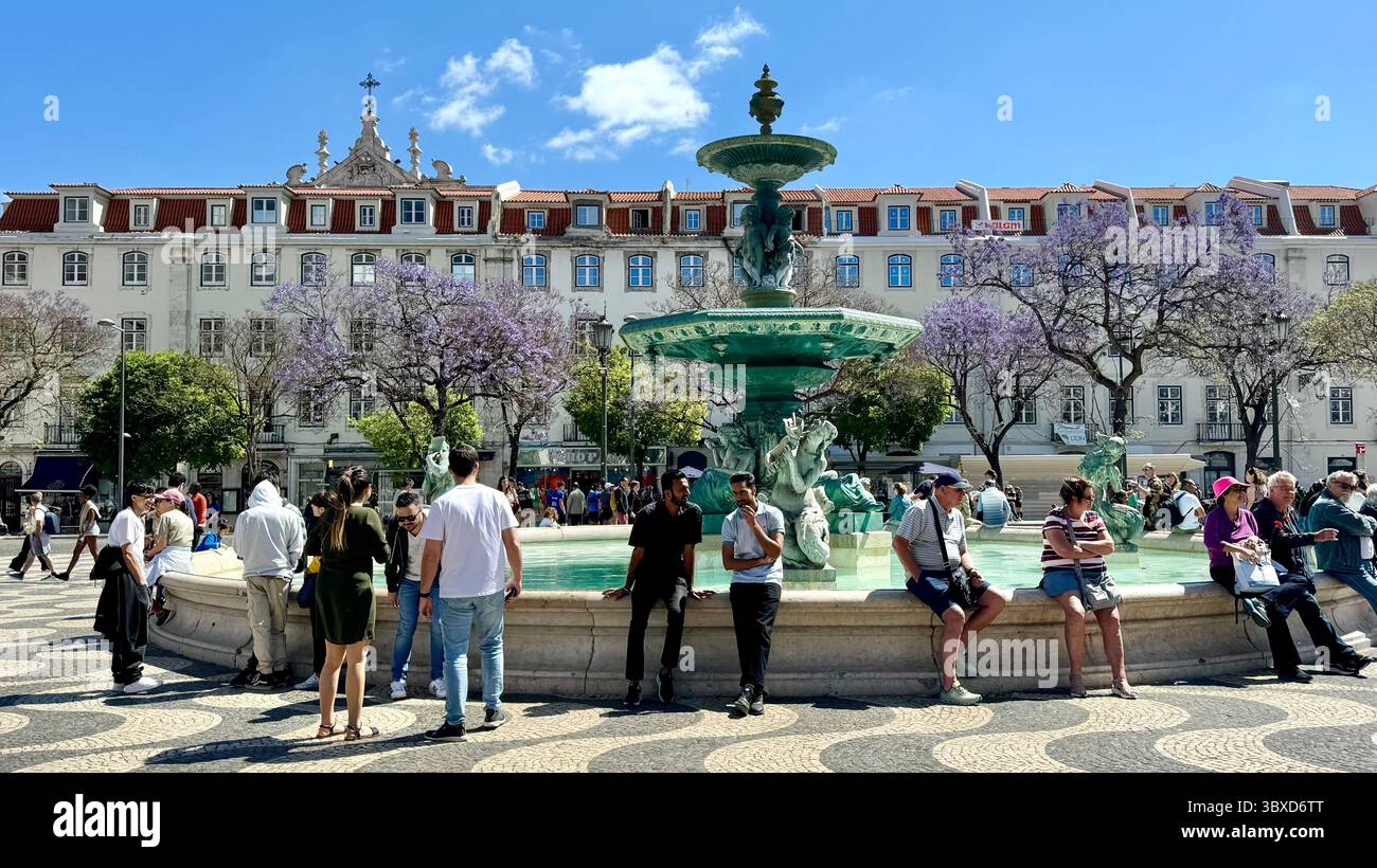 People sitting and standing around a fountain and flowering jacaranda trees in the Praco do Rossio, Lisbon Portugal - Smartphone Captured Stock Image