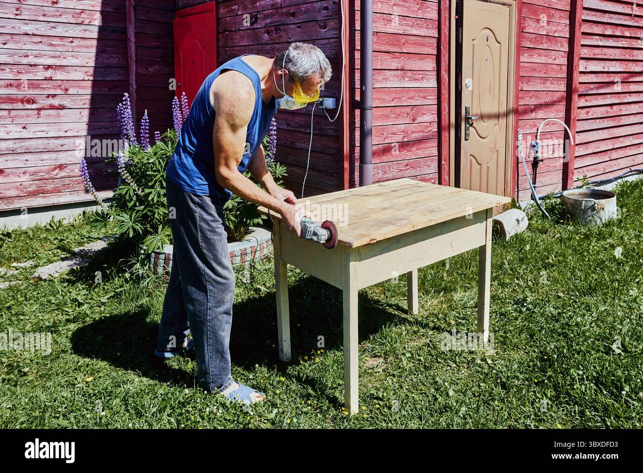 Working on tabletop edge with angle grinder, craftsman in protective gear sanding wood surface outside in rustic countryside setting Stock Photo