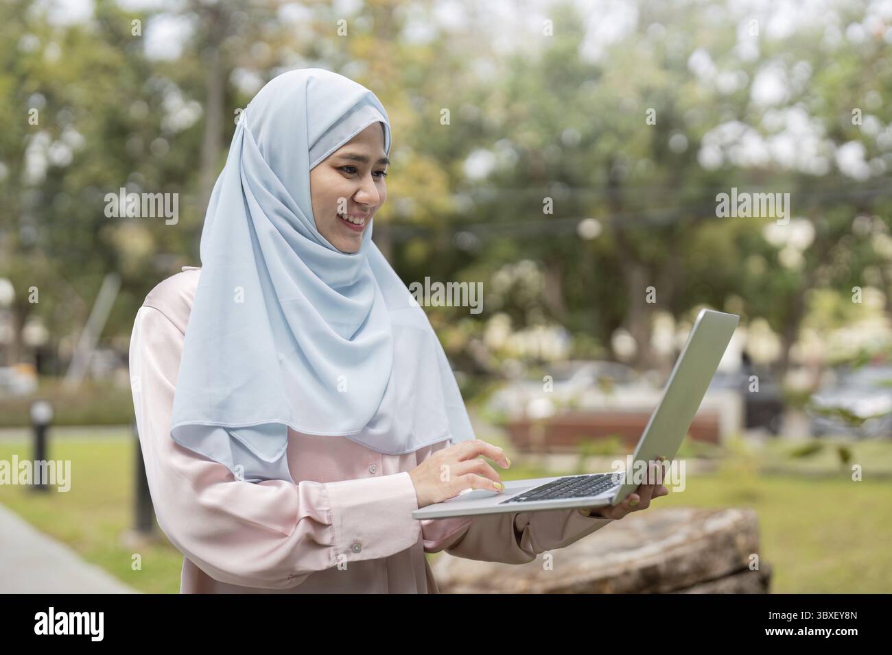 A confident Muslim woman in a hijab smiles as she types on her laptop ...