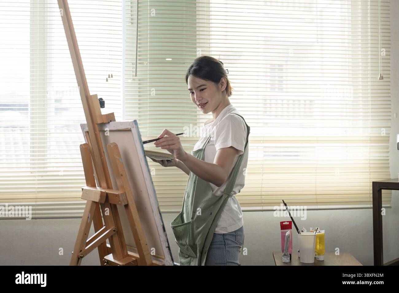 A young female artist is painting on a canvas in a bright workshop with ...