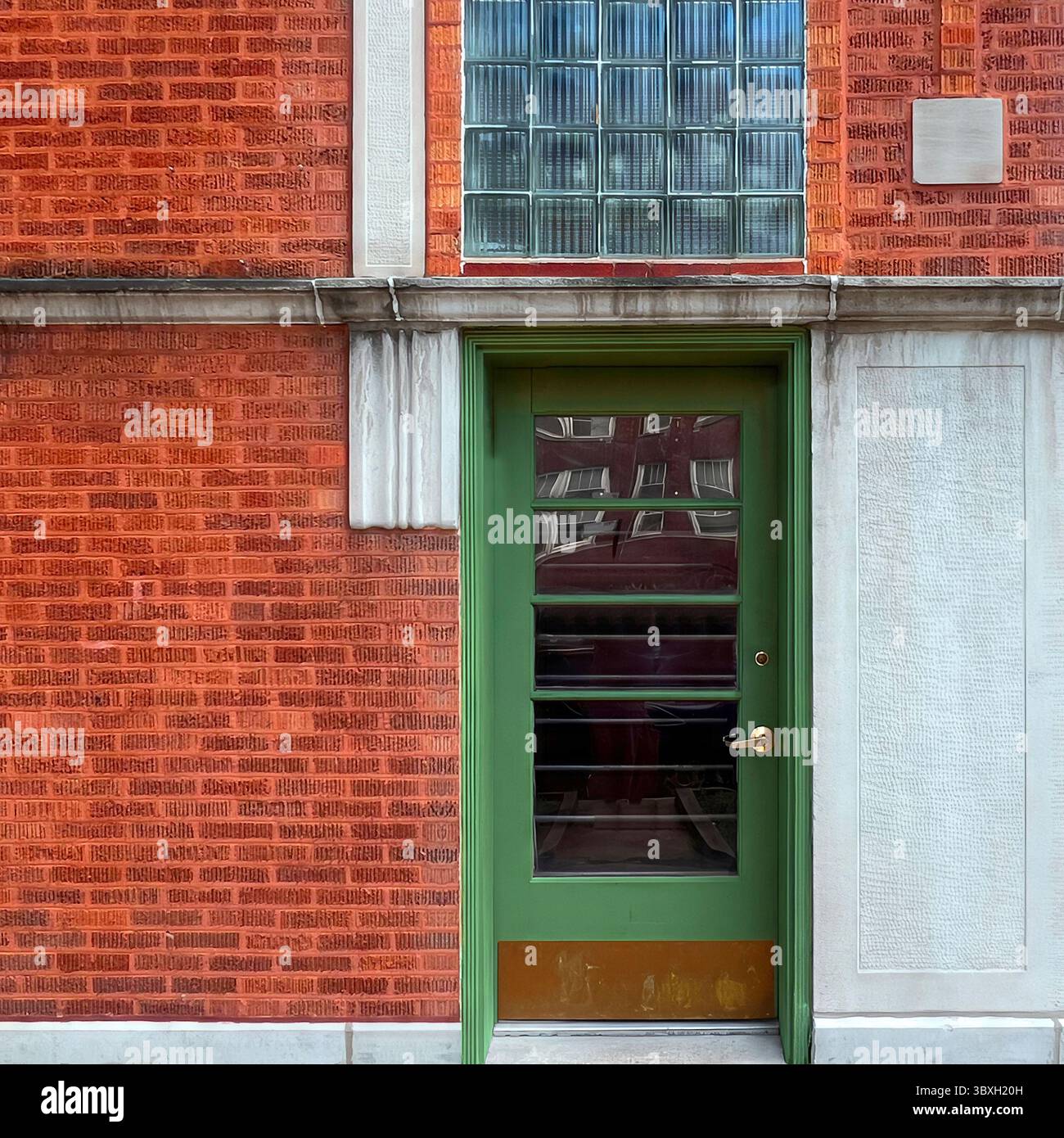 Modern green door with horizontal glass panes and brass kick plate on a textured red brick Chicago building, beneath a panel of vintage glass blocks. - Smartphone Captured Stock Image