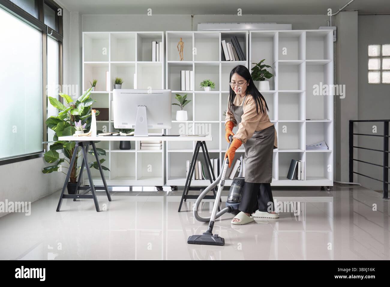 A professional maid or housekeeper cleaning a modern office space with a vacuum cleaner, ensuring a tidy and organized environment Stock Photo