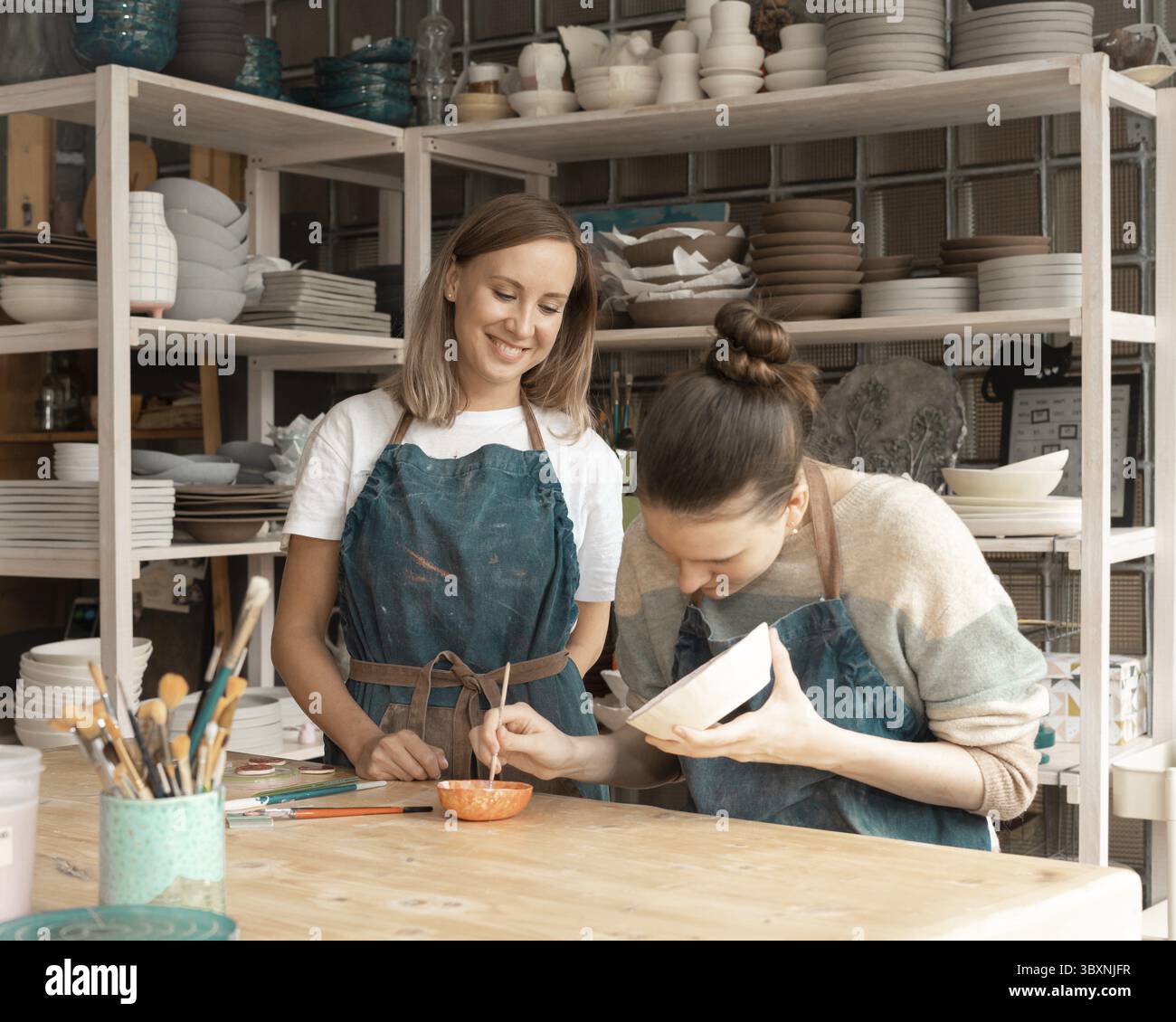 Woman making ceramic pottery. Attractive skilled young lady in apron standing at table and teaching student. Concept for workshop and master class Stock Photo
