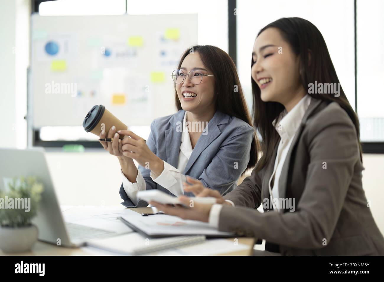 Two professional women in business attire working together in a bright, modern office, sharing ideas and smiling Stock Photo