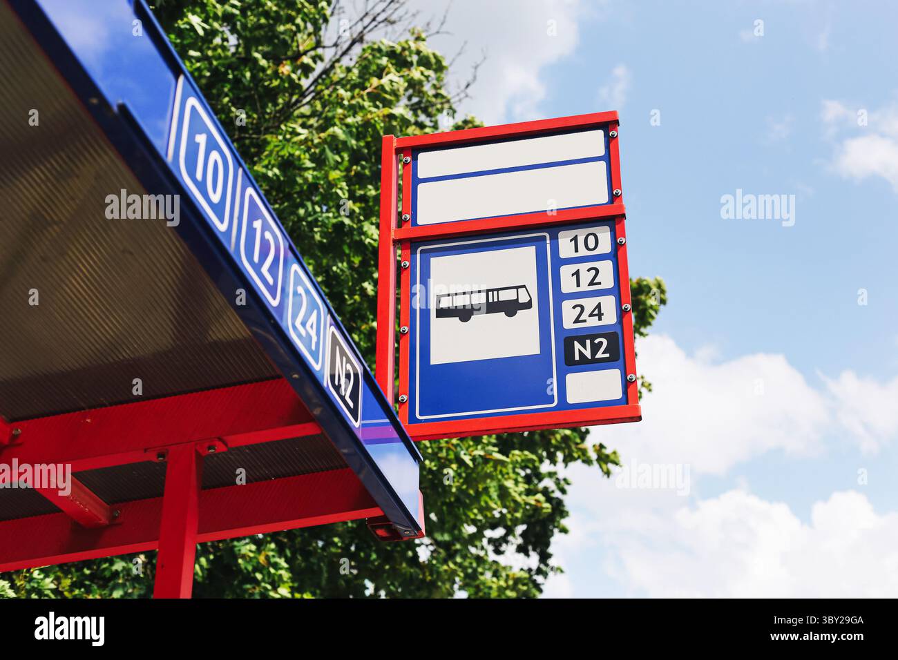 Urban bus stop signboard. Public transportation sign with timetable numbers. Red and blue bus station. Outdoor structure with tree and sky background. Stock Photo