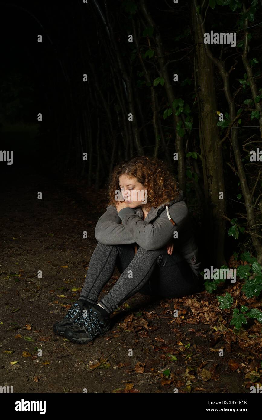 Woman sitting in dark forest, thoughtful and depressed Stock Photo