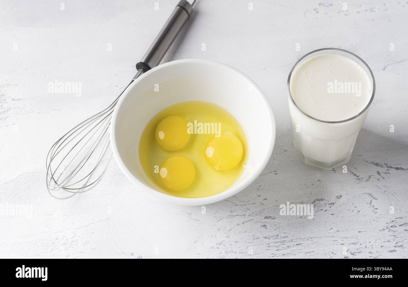 Food, White bowl with three broken eggs without shell, a glass of cream or milk and a whisk for whisking on a light gray background, top view. Cooking Stock Photo