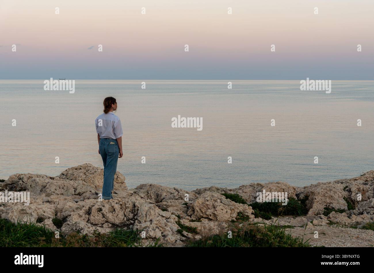Young woman stands on a rocky coast, gazing out at the tranquil ocean during a serene sunset Stock Photo