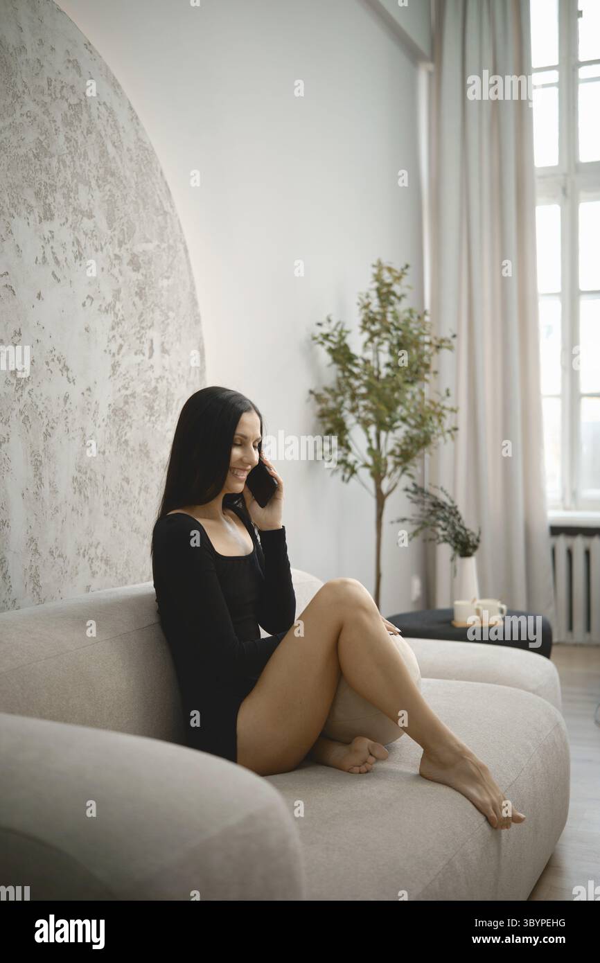 A young woman dressed in a black outfit sits comfortably on a light-colored couch Stock Photo