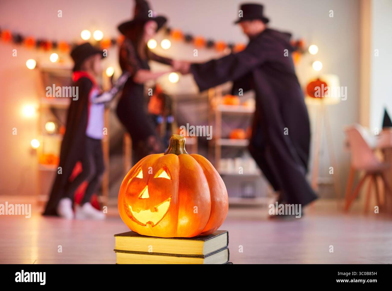 Glowing carved pumpkin standing on books in festive room while family in Halloween costumes dancing. Stock Photo
