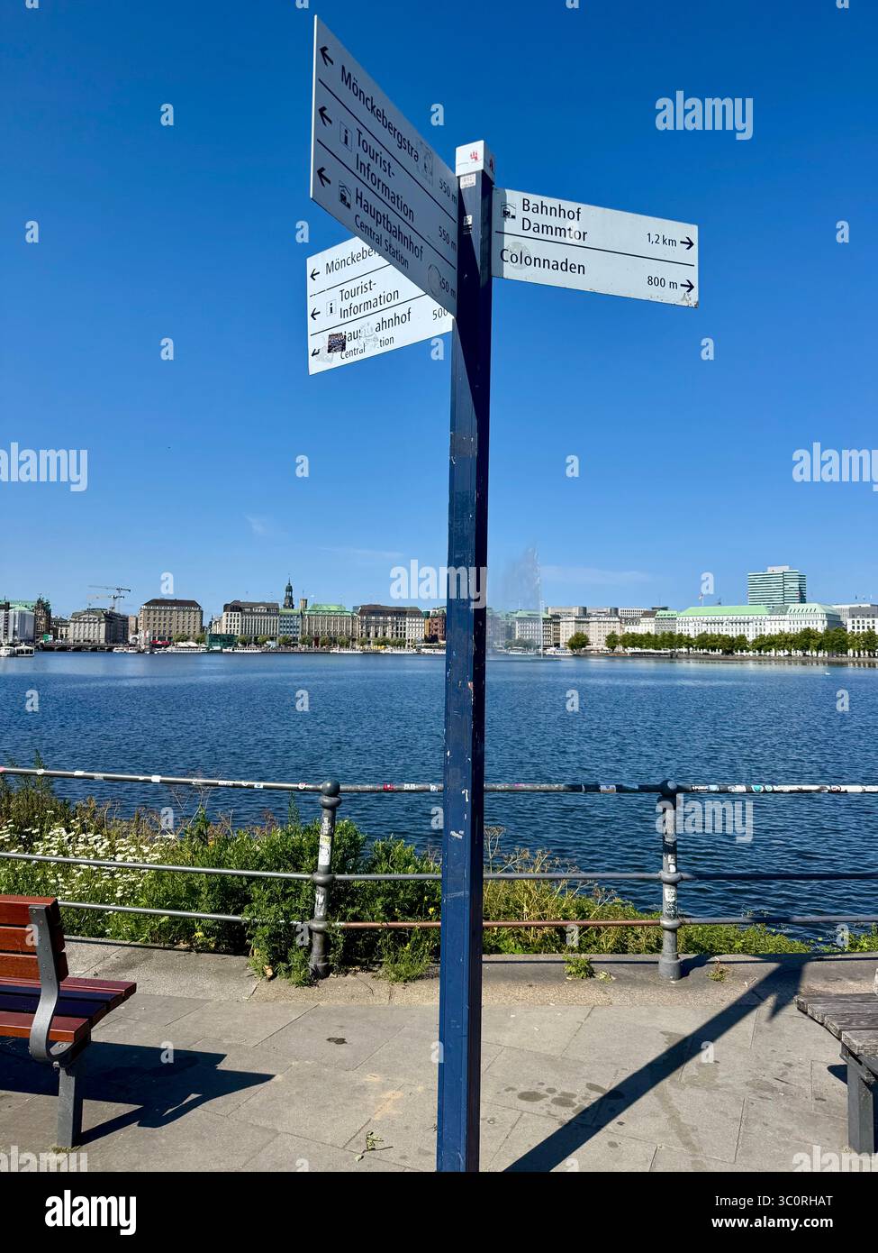 Directional signpost by the Inner Alster Lake (Binnenalster) with Hamburg cityscape in the background on a sunny day. Stock Photo