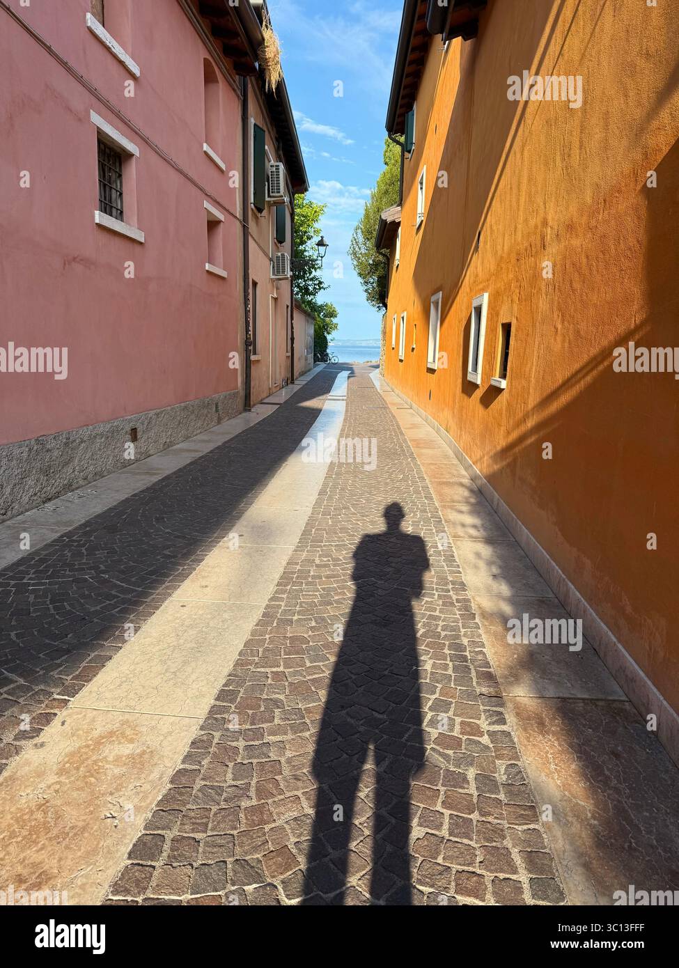 Photographer casting long shadow in the morning between colourful houses on an alleyway at Cisano leading to Lake Garda, Italy - Smartphone Captured Stock Image