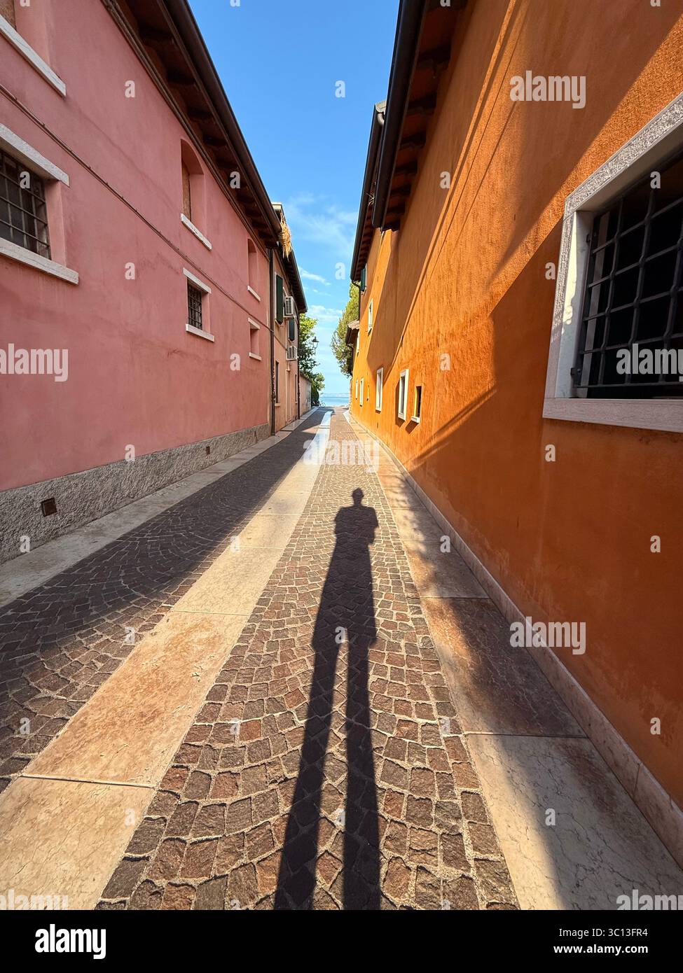 Photographer casting long shadow in the morning between colourful houses on an alleyway at Cisano leading to Lake Garda, Italy - Smartphone Captured Stock Image