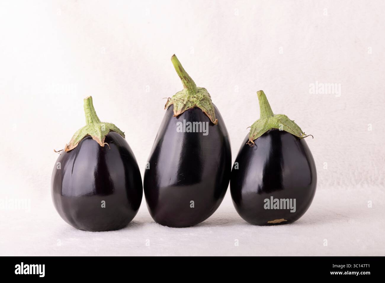 Three black eggplants are sitting on a white background. The eggplants are all different sizes and are arranged in a row Stock Photo