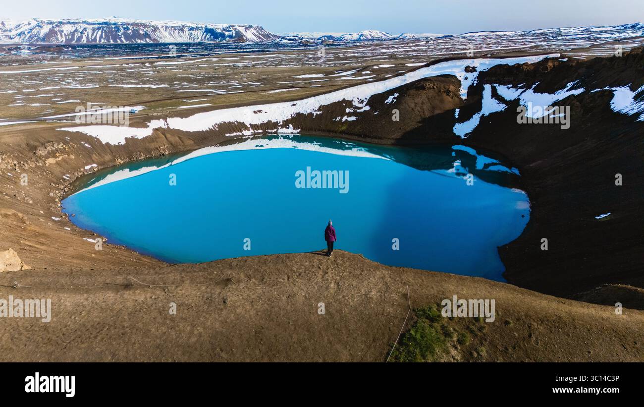 Nestled in the volcanic landscape of Iceland, Krafla crater presents a stunning view of a luminous blue lake surrounded by rugged terrain and snow-capped formations. Stock Photo